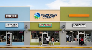 Edificio con fachada azul, beige, verde y gris; letreros "COFFEE", "AFROISA AUTENIING", logo "MIAMI-DADE COUNTY", "ICE CREAM", "STATIONERY". Personas en entradas.