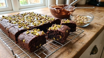 Primer plano de brownies de chocolate con trozos de pistacho espolvoreados encima, sobre una rejilla metálica. Hay cuencos con chocolate derretido y pistachos al fondo.