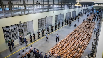 Filas de reclusos con el torso descubierto sentados en el suelo dentro de una prisión, custodiados por guardias de seguridad armados y uniformados