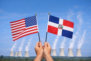 Dos manos sostienen banderas de Estados Unidos y República Dominicana. Al fondo, torres de enfriamiento de una planta nuclear emiten vapor bajo un cielo azul.