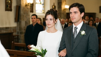 Una novia con vestido blanco, velo y ramo de calas, camina del brazo de un novio con traje gris y ramillete en una iglesia con bancos de madera y vidrieras