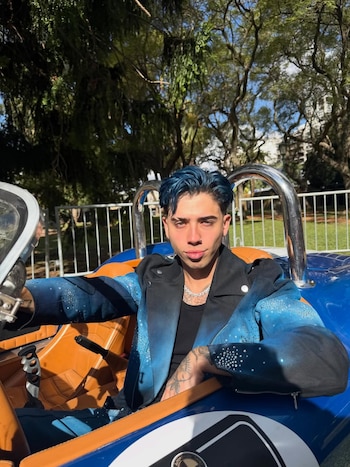 Hombre joven con cabello azul brillante, chaqueta azul y negra con pedrería, y camiseta negra, sentado en un auto deportivo azul y naranja