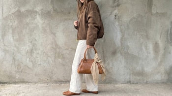 Vista lateral de una persona con chaqueta de ante marrón, pantalones blancos y mocasines. Sostiene un bolso de cuero con un pañuelo de lunares, frente a una pared gris.