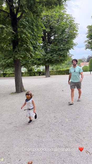 Hombre adulto y niño pequeño caminan por un camino de grava en un parque con árboles y setos. El cielo está parcialmente nublado