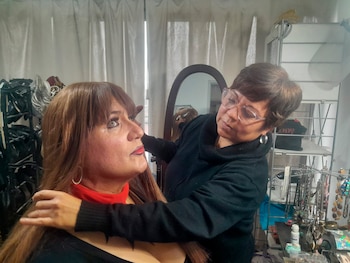 Una persona con peluca castaña, maquillaje y pañuelo rojo es asistida por otra persona que ajusta su cabello en un estudio con cortinas y estantes