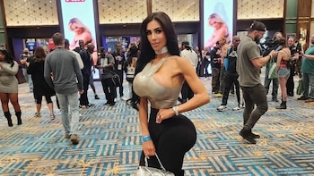 Una mujer con cabello largo oscuro, un top asimétrico brillante y pantalones negros, posa en un salón concurrido