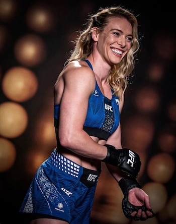 Luchadora de UFC con cabello rubio rizado, top y shorts azules con el logo UFC, guantes negros de MMA, sonriendo con un fondo bokeh oscuro
