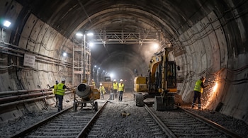 Interior de un túnel ferroviario con vías, grava, una excavadora, una mezcladora y cinco trabajadores con cascos y chalecos amarillos; uno corta metal con chispas.