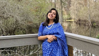Mujer joven con sari azul y collar de perlas blancas sonríe, apoyada en una baranda de madera con un fondo de lago y árboles