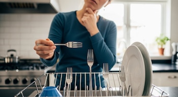 Una mujer en la cocina sostiene un tenedor sobre un escurridor con otros cubiertos y platos, mientras se lleva la mano a la barbilla pensativa.