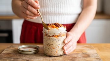 Frasco de vidrio con avena, yogur, semillas de chía, zanahoria y nueces sobre una tabla de madera, sostenido por una persona con una cuchara dorada.