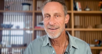 Un hombre de mediana edad con cabello corto y barba gris, camiseta verde claro y collar oscuro, sonríe a la cámara. Detrás hay una estantería de madera con objetos