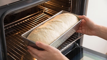 Manos introducen un molde rectangular de metal con masa de pan cruda sobre la rejilla de un horno eléctrico encendido con luz interior.