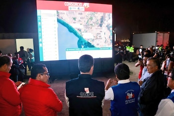 Vista trasera de un grupo de personas, algunos con chalecos de ONPE y Defensoría del Pueblo, observando atentamente una gran pantalla que muestra un mapa satelital