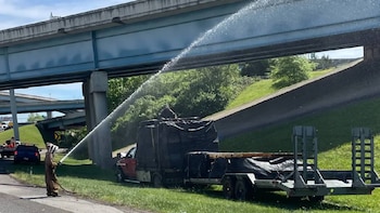 Un trabajador con traje protector beige y blanco rocía un chorro de agua sobre un camión rojo y su remolque con lona negra bajo un paso elevado de la I-40