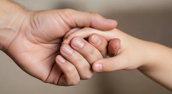 Primer plano de una mano de adulto grande y una mano de niño pequeña entrelazadas, con un fondo claro y desenfocado.