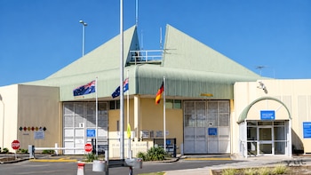 Edificio con techo verde angular, dos portones enrejados, cabina de entrada, barreras vehiculares y banderas de Australia y del pueblo aborigen