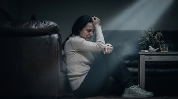 Mujer con jersey claro y pantalones oscuros sentada en el suelo con lágrimas en el rostro junto a una mesa auxiliar y un sofá de cuero.