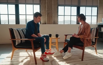 Sam Altman y una mujer sentados en sillones de madera clara, conversando. Entre ellos hay una mesa con dos tazas negras y grandes ventanales de fondo