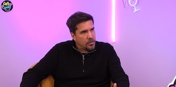Hombre de cabello oscuro y barba, vestido con suéter negro, sentado en un sillón amarillo frente a un fondo rosa con luces de neón y un logo