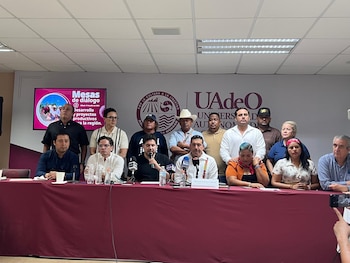 Un grupo de dieciseis personas, hombres y mujeres, sentadas y de pie en una sala, con una mesa delante que tiene micrófonos y botellas de agua. Hay un monitor con texto y un logo