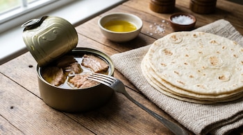 Lata de atún abierta con tenedor, atún en aceite, tortillas de trigo apiladas sobre paño, cuencos con aceite y sal, todo en superficie de madera.