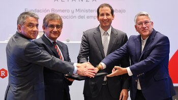 Cuatro hombres con traje y corbata colocan sus manos juntas sobre una carpeta negra durante un evento institucional