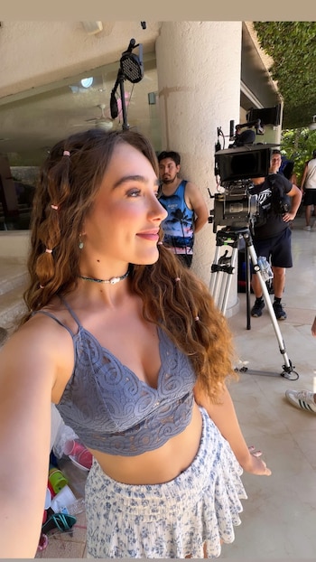 Una mujer joven con cabello largo y ondulado, top de encaje azul y falda floral, sonríe en un primer plano. Equipo de filmación y personal al fondo