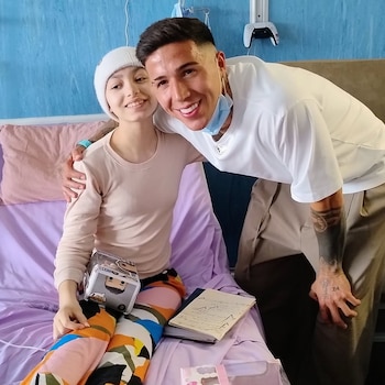 Enzo Fernández sonríe junto a una paciente con gorro blanco sentada en una cama de hospital sosteniendo una caja de juguete
