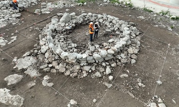 Dos personas con chalecos reflectantes observan una estructura circular de piedras en un terreno delimitado por una red de hilos