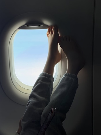 Primer plano de los pies desnudos de una niña, apoyados en el marco superior de una ventana de avión con el cielo azul de fondo