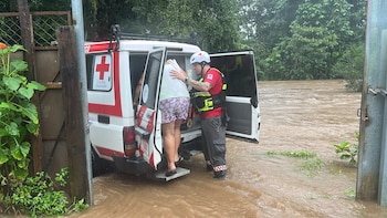 Tres rescatistas con uniformes de la Cruz Roja, cascos y chalecos, junto a un vehículo de rescate blanco. El fondo muestra un paisaje montañoso y húmedo con vegetación