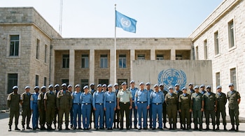 Grupo de 21 efectivos uniformados de diversas etnias posa frente a un edificio institucional con la bandera azul de la ONU y su logo en la pared.