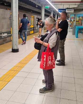 Mujer mayor con cabello gris y un bolso rojo mirando su teléfono móvil en una estación de metro con un tren y otras personas de fondo
