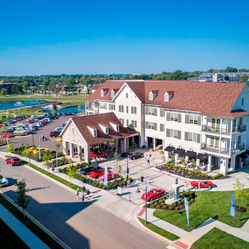 Toma aérea de The Inn at Meadowbrook Hotel, un edificio blanco con techo marrón, rodeado de una plaza y estacionamiento lleno de coches deportivos y personas
