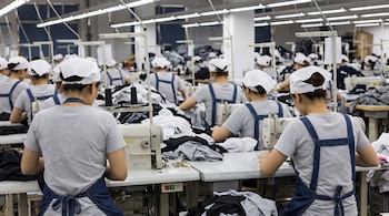 Filas de trabajadores con gorros blancos y delantales azules operan máquinas de coser industriales en un taller con mesas y telas.