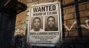 Un cartel de "SE BUSCA" con fotos de dos hombres y una recompensa de $10,000 está pegado con cinta adhesiva a una pared de ladrillos con grafitis, parcialmente iluminada por el sol.