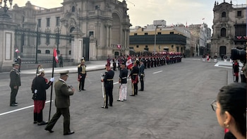 Oficiales y militares con uniformes de gala y banderas peruanas forman una línea en una calle frente a un edificio histórico grande con rejas, bajo un cielo nublado