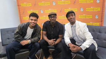 Tres hombres sentados en un sofá negro frente a un fondo amarillo y naranja con logotipos del musical Buena Vista Social Club