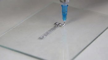 Primer plano de una punta de jeringa azul y gris imprimiendo una estructura metálica plateada de tres brazos sobre una superficie de vidrio transparente.