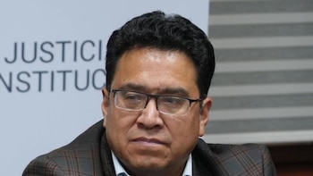 Primer plano de un hombre con gafas y traje a cuadros, frente a un cartel borroso con las palabras 'Justicia' y 'Institucional'