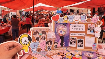 Vista cercana de un puesto en un tianguis con toldos rojos, exhibiendo freebies de K-Pop de BTS: stickers de personajes BT21, caricaturas de miembros y galletas. Una mano sostiene un sticker.