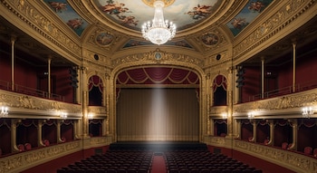 Un teatro vacío con butacas rojas, escenario con telón beige, palcos dorados, araña de cristal en el centro y frescos en el techo.
