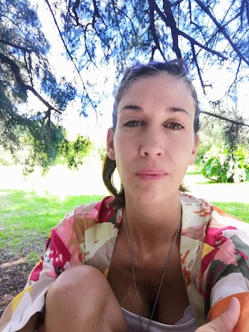 Una mujer de cabello recogido y prenda estampada posa al aire libre frente a varios árboles y un área con césped