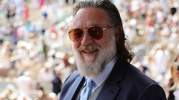 Russell Crowe sonríe mientras sostiene una pelota de tenis amarilla con el logo de Roland Garros, vistiendo traje y gafas de sol, con el público de fondo