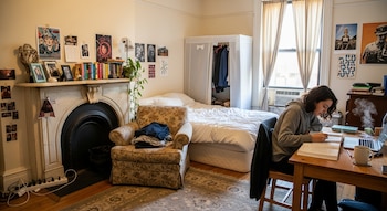Una joven estudia en un escritorio con libro y portátil. La habitación tiene una cama, sillón con ropa, armario, chimenea decorativa y ventana con cortinas.