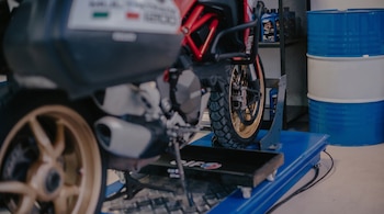 Dos motocicletas sobre un elevador azul. Una motocicleta tiene chasis rojo y ruedas doradas. Un estante con botellas y barriles azules y blancos al fondo.