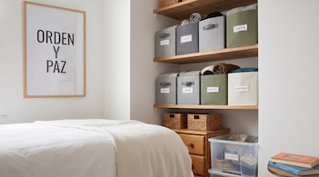 Estantes de madera con cajas de tela etiquetadas, canastas de mimbre, una cama con ropa blanca y un cuadro con texto en la pared.