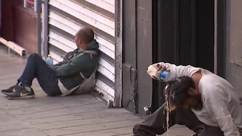 Dos hombres sentados en una acera gris. Uno, con barba y camiseta blanca, vierte líquido sobre su cabeza. El otro, calvo con sudadera verde, se apoya en una pared