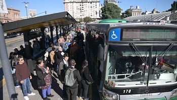 Una multitud de personas espera en una parada de autobús en una calle urbana. Un autobús gris está parado en la vía, y edificios se ven al fondo bajo un cielo azul